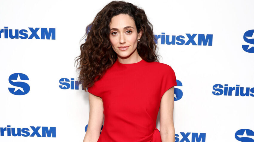 Emmy Rossum wears a red T-shirt while standing in front of a white SiriusXM backdrop.