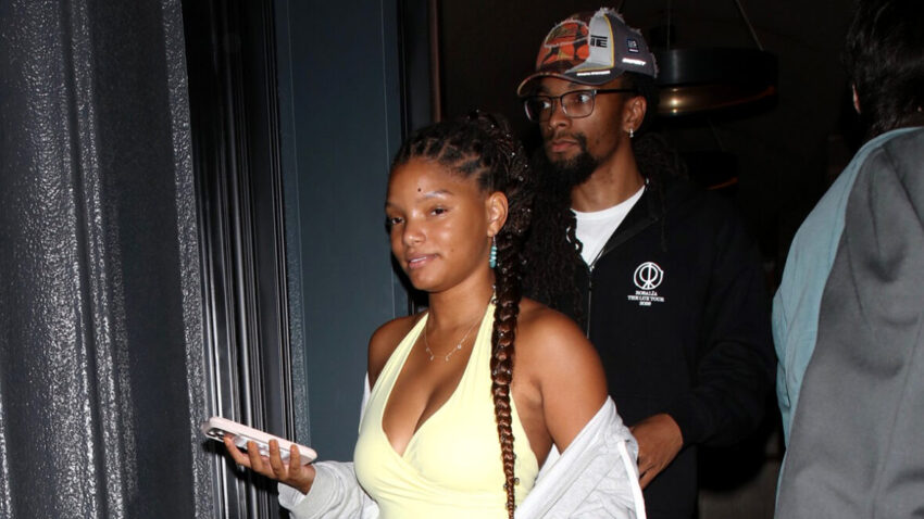 Halle Bailey in a yellow jumpsuit walking out of a restaurant with her phone in hand. Scott Bridgeway seen walking out behind her in a black hoodie.