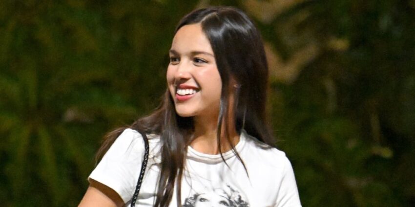 Olivia Rodrigo in a white t-shirt against some greenery in the background, smiling and looking to the left